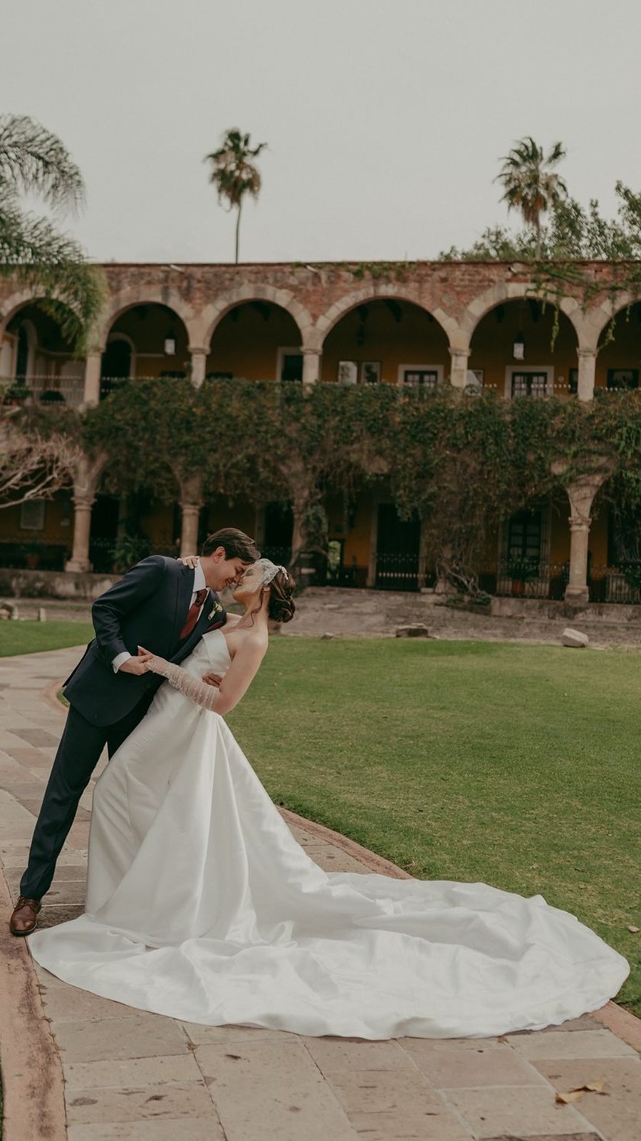 Novio inclina a la novia para besarla frente a una hacienda con arcos y palmeras