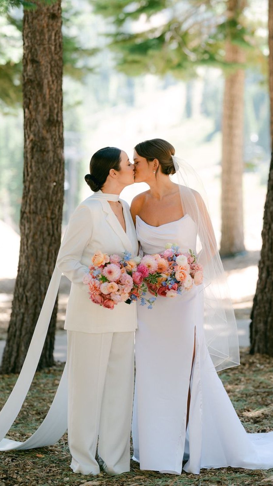 Dos novias se besan en un bosque, una con traje blanco y otra con vestido, ambas con ramos de colores