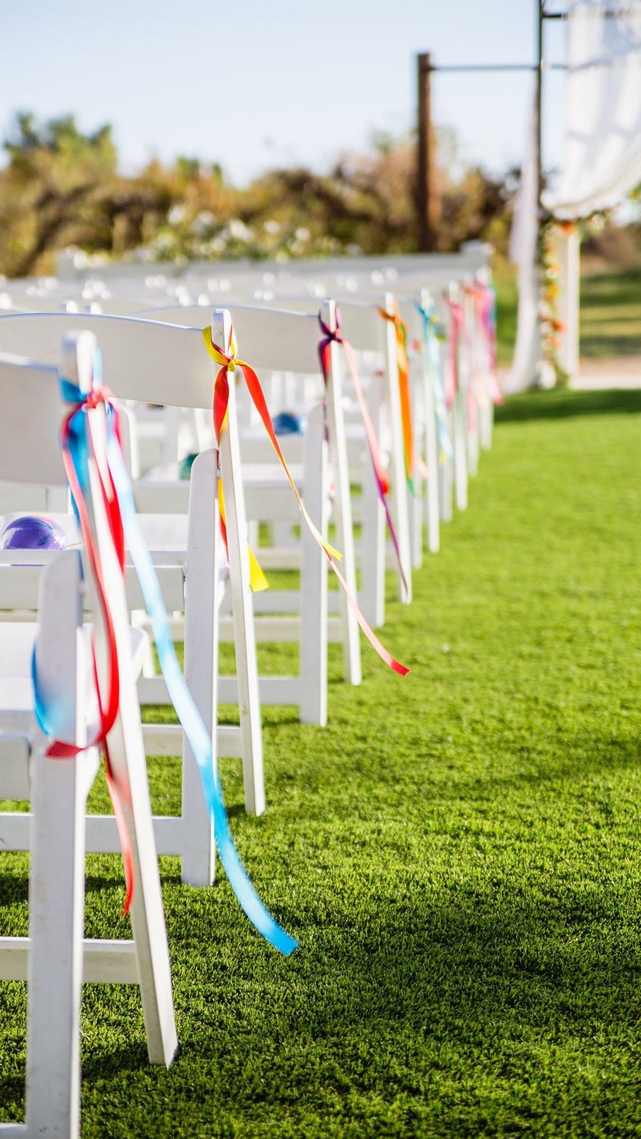 Sillas de ceremonia decoradas con listones de colores del arcoíris en un jardín
