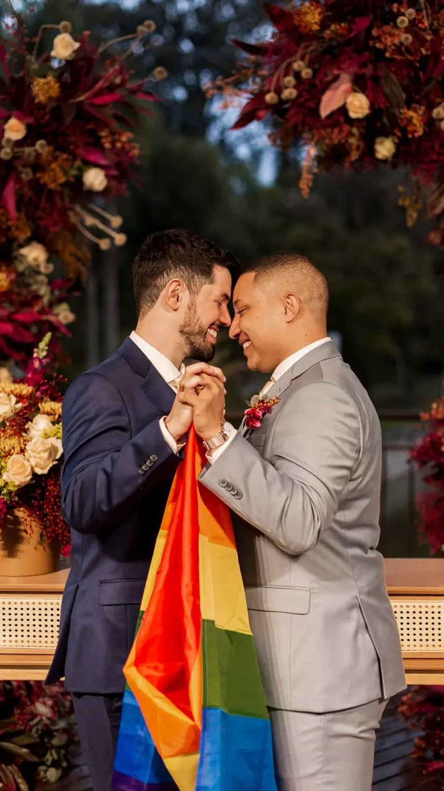 Dos novios con trajes azul y gris sostienen una bandera arcoíris durante su boda