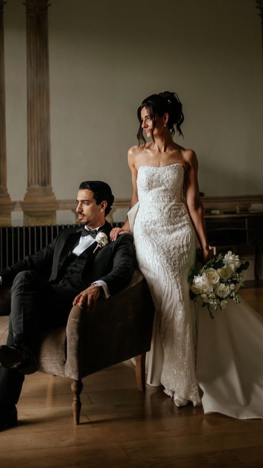 Novios posan en un salón con piso de madera, ella de pie con vestido de encaje