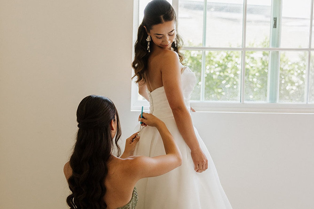 Dama de honor abrocha el vestido de la novia junto a una ventana