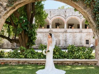 Novia en el jardín de una hacienda enmarcada por un arco de piedra con arcadas al fondo