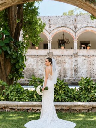 Novia en el jardín de una hacienda enmarcada por un arco de piedra con arcadas al fondo