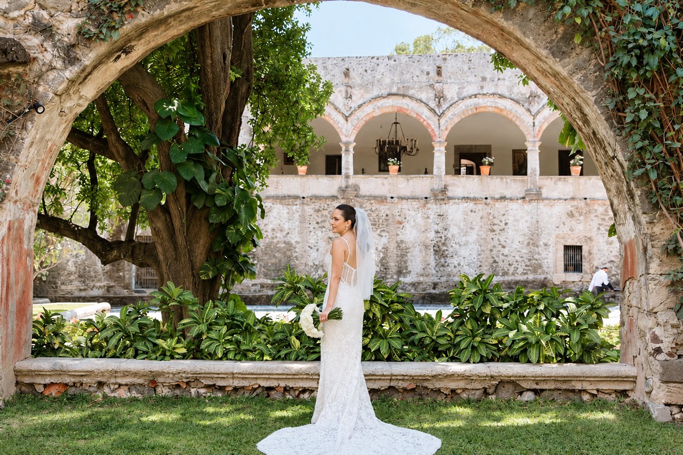 Novia en el jardín de una hacienda enmarcada por un arco de piedra con arcadas al fondo