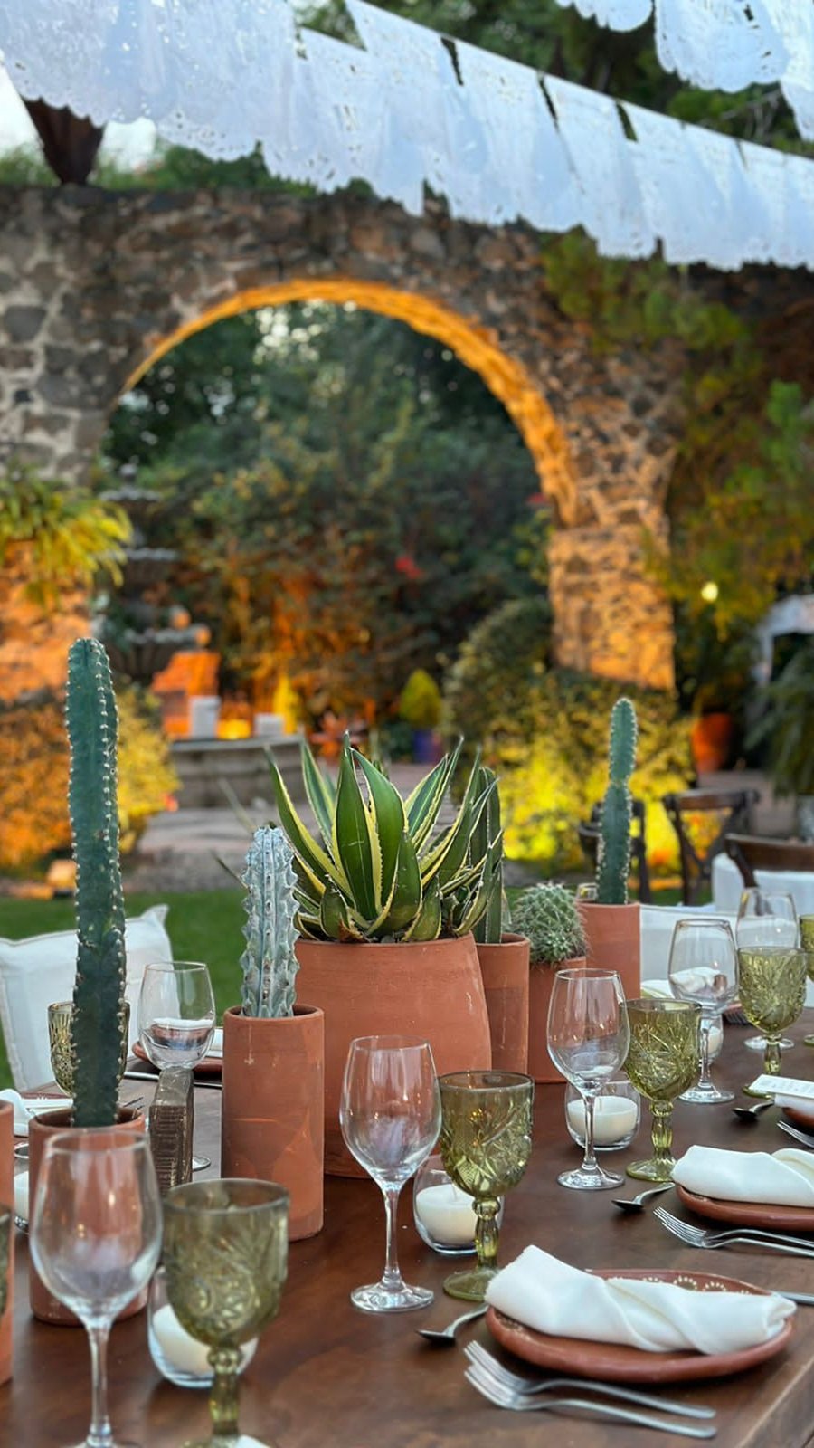 Mesa con cactus, agaves y cristalería verde bajo un arco de piedra con papel picado