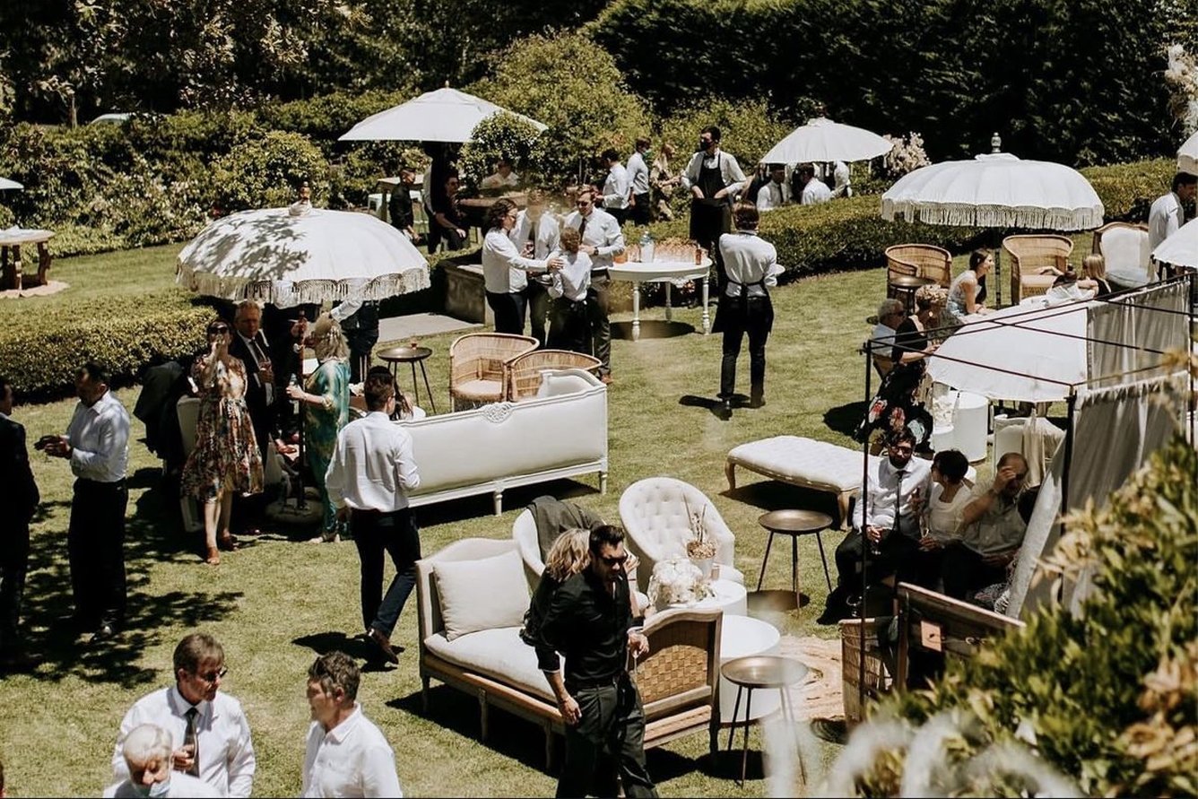 Invitados conviven en un jardín con salas lounge y sombrillas durante una tornaboda